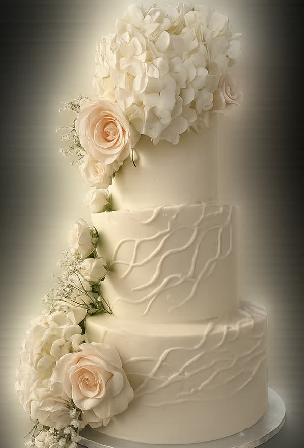 Three-tier round white buttercream wedding cake with piped vine texture on the lower two tiers, a hydrangea cluster on top and a cascade of blush roses and baby's breath down the left side.