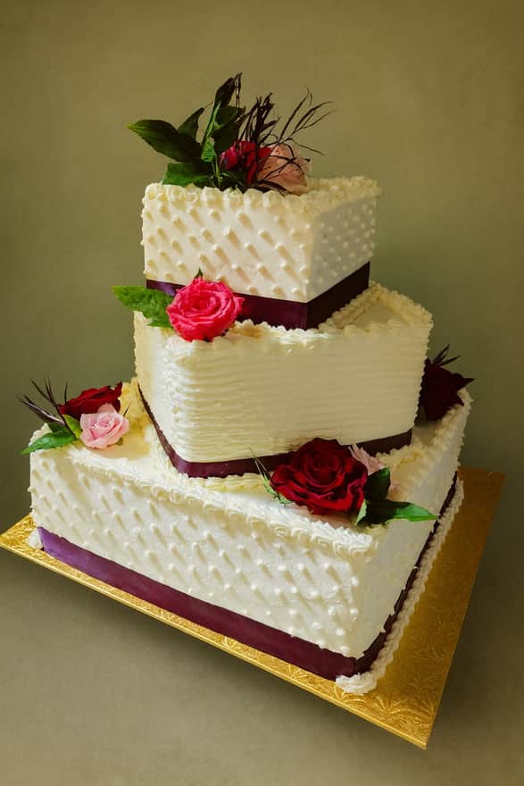 Three-tier square white buttercream wedding cake with a quilted dot pattern on the top and bottom tiers and a combed middle tier, each banded with a deep aubergine ribbon and set with fresh red and pink roses.
