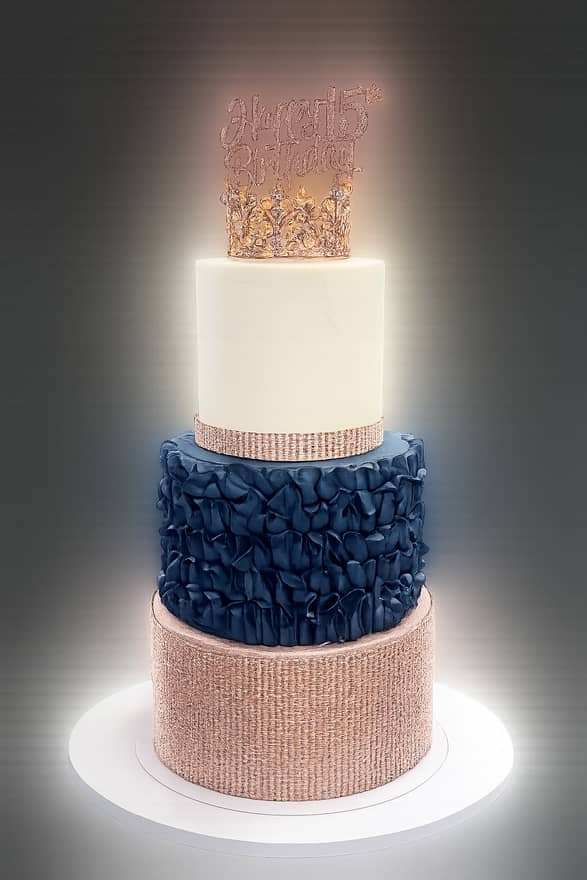 Three-tier round cake with a rose-gold textured bottom tier, a navy blue ruffled middle tier and a smooth ivory top tier, topped with a gold rhinestone crown and a glittered birthday script topper.