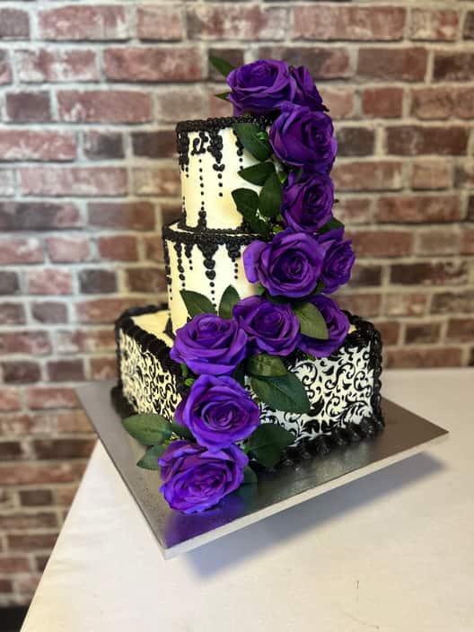 Three-tier wedding cake with a square ivory base tier stencilled in black damask and two round upper tiers piped with black beaded swags, trimmed with a cascade of deep purple roses and green leaves.
