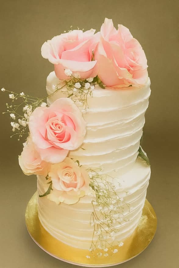 Two-tier round white wedding cake in horizontally combed buttercream, dressed with fresh pale pink and cream roses and sprigs of baby's breath cascading down one side, on a gold cake board.