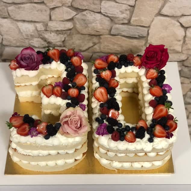 Number cake shaped as a five and a zero, built from layers of pale sponge and piped white cream, topped with glazed strawberries, blackberries, blueberries, raspberries and pink and burgundy roses.