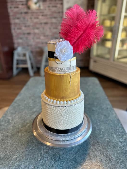 Four-tier art deco cake with a gold-leaf tier, an ivory tier piped with feather-fan patterns, black satin ribbons, strands of silver pearls and a white sugar rose, topped with a tall crimson ostrich feather.
