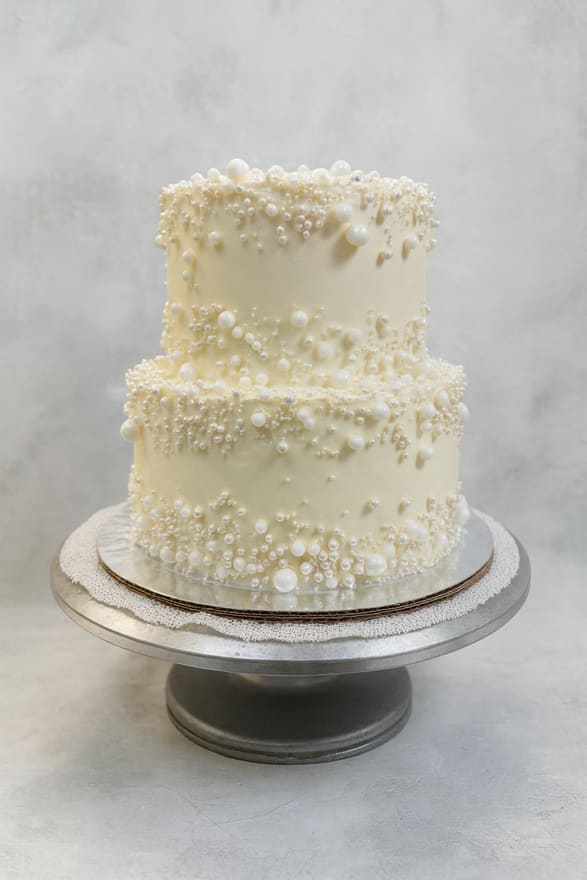 Two-tier round ivory buttercream cake covered in graduated white and silver sugar pearls, set on a lace-lined pewter cake stand.