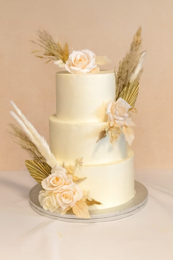 Three-tier round ivory buttercream wedding cake decorated with cream roses, dried palm fans and tall pampas grass plumes, on a silver board over a white cloth.