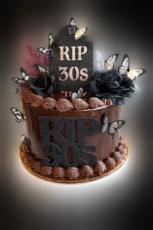 Single-tier cake covered in glossy chocolate ganache with piped chocolate rosettes top and bottom, a black glitter &lsquo;RIP 30s&rsquo; headstone topper, dressed with black roses and cream-and-black paper butterflies.