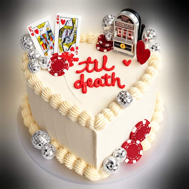 Heart-shaped cake iced in ivory buttercream with a shell-piped border and &lsquo;til death&rsquo; written in red piping across the top, decorated with king and queen of hearts playing cards, red poker chips and a miniature slot machine.