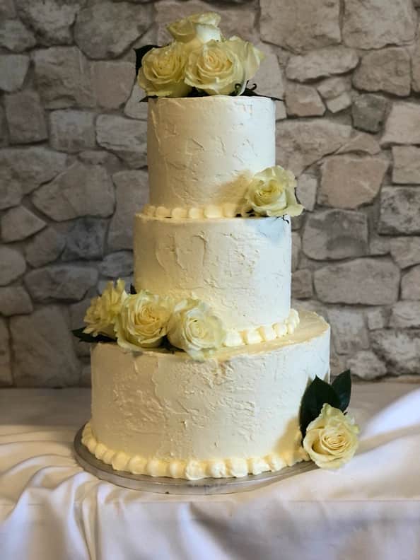 Three-tier round wedding cake in ivory buttercream with a rustic palette-knife finish, pearl piping between the tiers, and clusters of cream roses with dark green leaves cascading down the front.