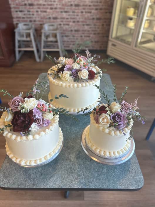 Three single-tier ivory buttercream cakes with piped shell borders, each crowned with clusters of mauve, cream and deep burgundy roses and eucalyptus.