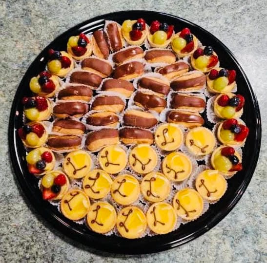 Round black catering tray filled in concentric rings with chocolate-glazed mini éclairs at the centre, lemon curd tartlettes each piped with a chocolate flourish, and fresh fruit tartlettes topped with strawberry, blueberry and green grape.