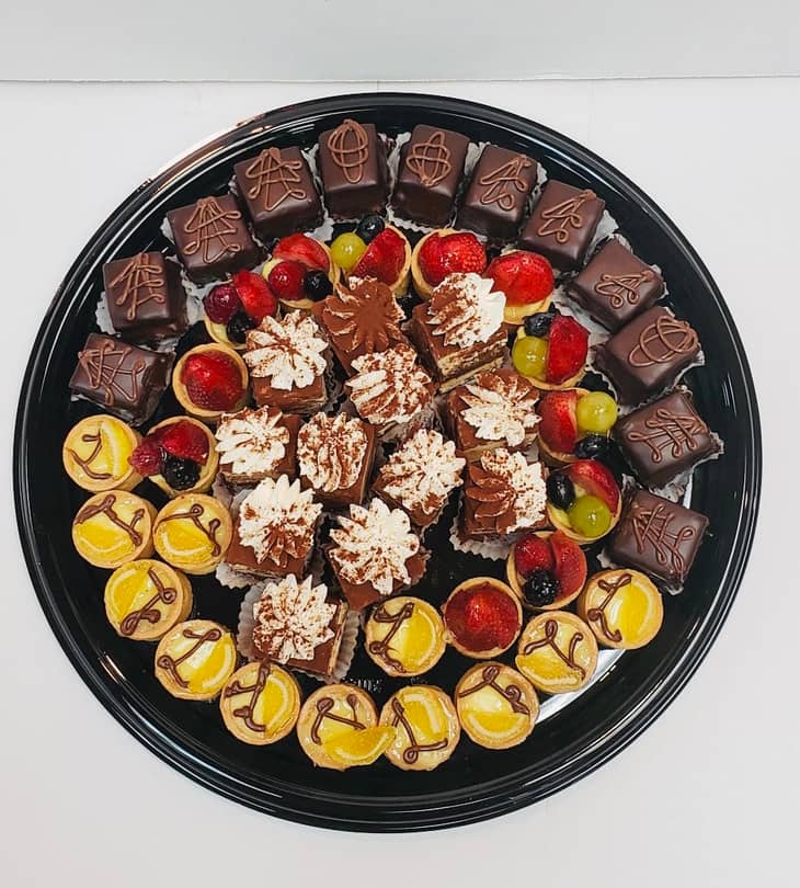 Round black catering tray photographed from above, ringed with chocolate-enrobed petit fours piped with fine chocolate scrollwork, cocoa-dusted tiramisu squares and mille-feuille slices at the centre, lemon tartlettes and fresh fruit tartlettes around them.