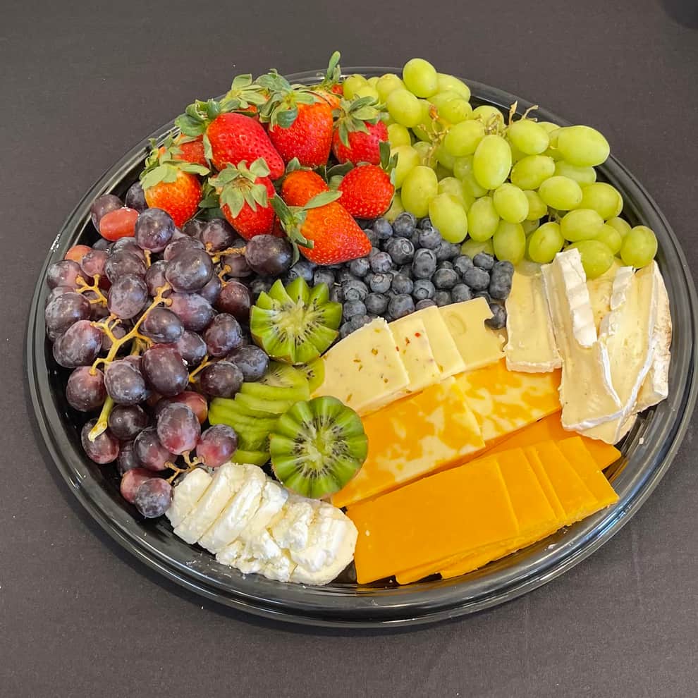 Round black catering tray arranged in wedges with strawberries, green and red grapes, blueberries and sliced kiwi on one side and sliced cheddar, marbled cheese, pepper jack, brie wedges and crumbled fresh goat cheese on the other.