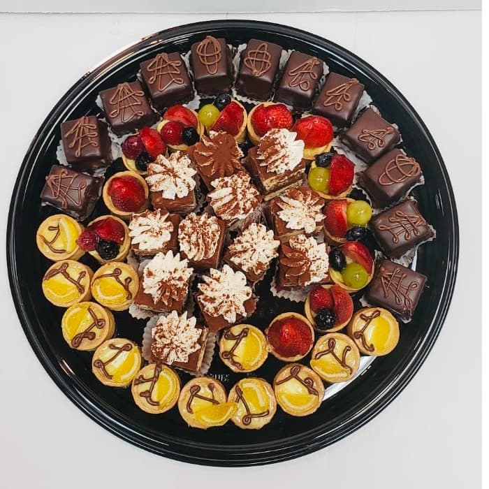Round black tray composed in rings: chocolate-glazed petit fours with piped scrollwork around the outside, fresh fruit tartelettes and lemon tartlettes in the middle band, and cocoa-dusted layered squares with cream rosettes at the centre.