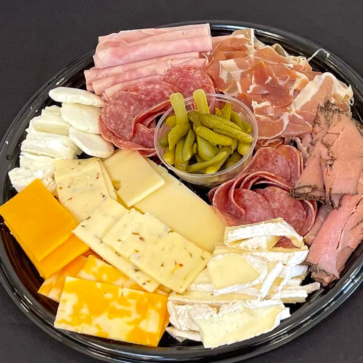 Round black catering tray holding fanned slices of ham, salami, prosciutto and rare roast beef beside cheddar, pepper jack, Swiss, brie and fresh goat cheese, with a cup of cornichons at the centre.