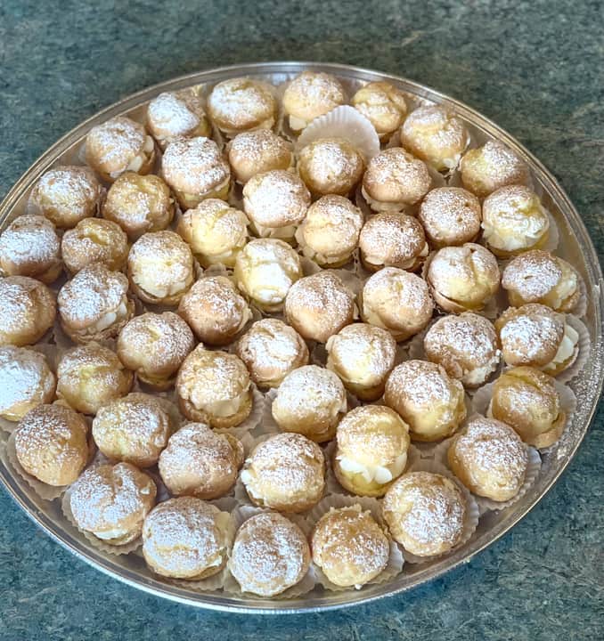 Overhead view of a round silver tray packed with small golden cream puffs in paper cups, split and filled with white cream and dusted with powdered sugar.