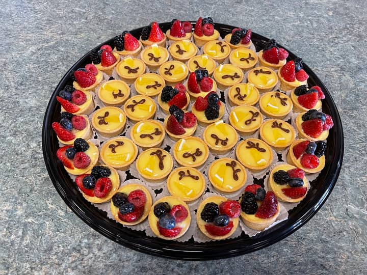 Large round black tray filled with mini tartlettes: an outer ring topped with strawberries, raspberries, blackberries and blueberries surrounding rows of lemon tartlettes glazed yellow and finished with a chocolate flourish.
