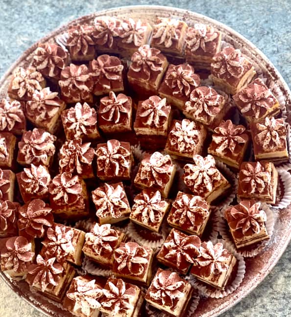 Overhead view of a round tray holding about fifty square mini tiramisu, layers of sponge and pale mascarpone cream visible at the sides, each topped with a piped cream rosette and a heavy dusting of cocoa powder.