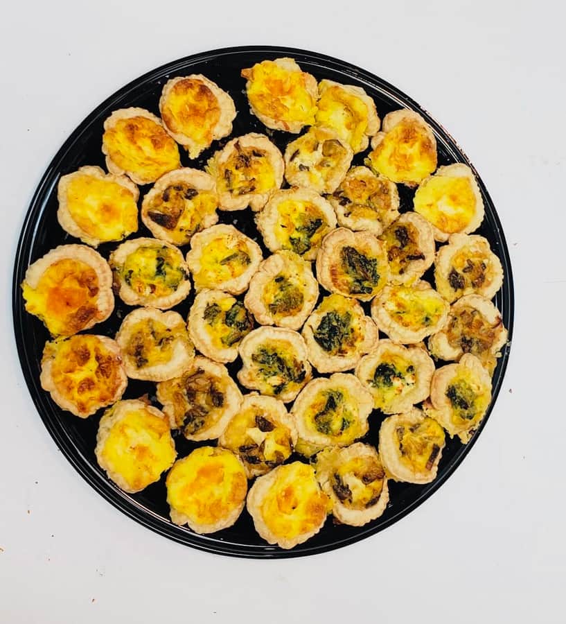 Round black catering tray photographed from above, packed with about forty miniature quiches in fluted all-butter pastry shells with golden custard fillings, some studded with spinach, some with mushroom and onion.