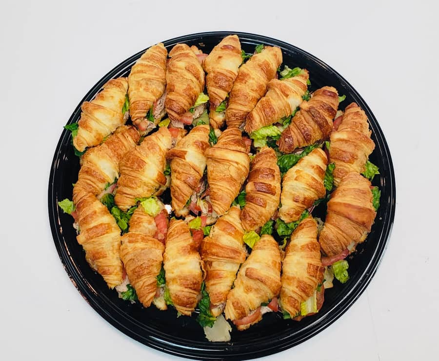 Overhead view of a round black catering tray filled with about twenty mini butter-croissant sandwiches, each split and filled with sliced ham or turkey, romaine lettuce and diced tomato.