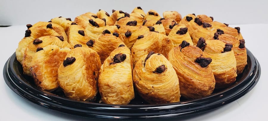 Eye-level view of a round black tray tightly packed with mini pain au chocolat, golden laminated layers visible in cross-section, each topped with a piece of dark chocolate.