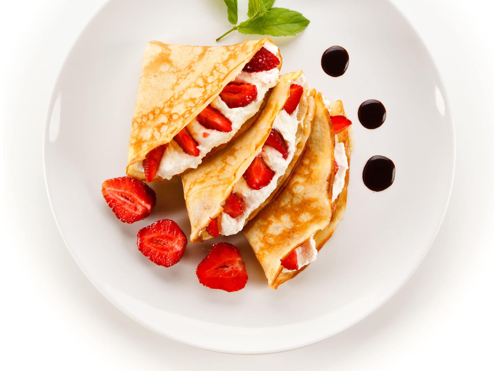 Folded French crepes filled with whipped cream and sliced strawberries on a white plate, with halved fresh strawberries beside them; the plate is garnished for serving with drops of chocolate sauce and a sprig of mint.