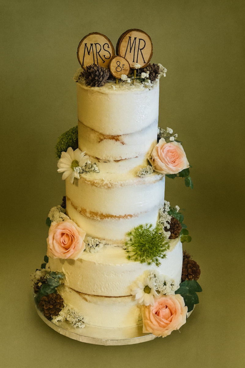 Three-tier semi-naked wedding cake, the sponge showing through a thin white coat, dressed with peach roses, white daisies, thistle, pinecones and moss under wooden MRS and MR toppers.