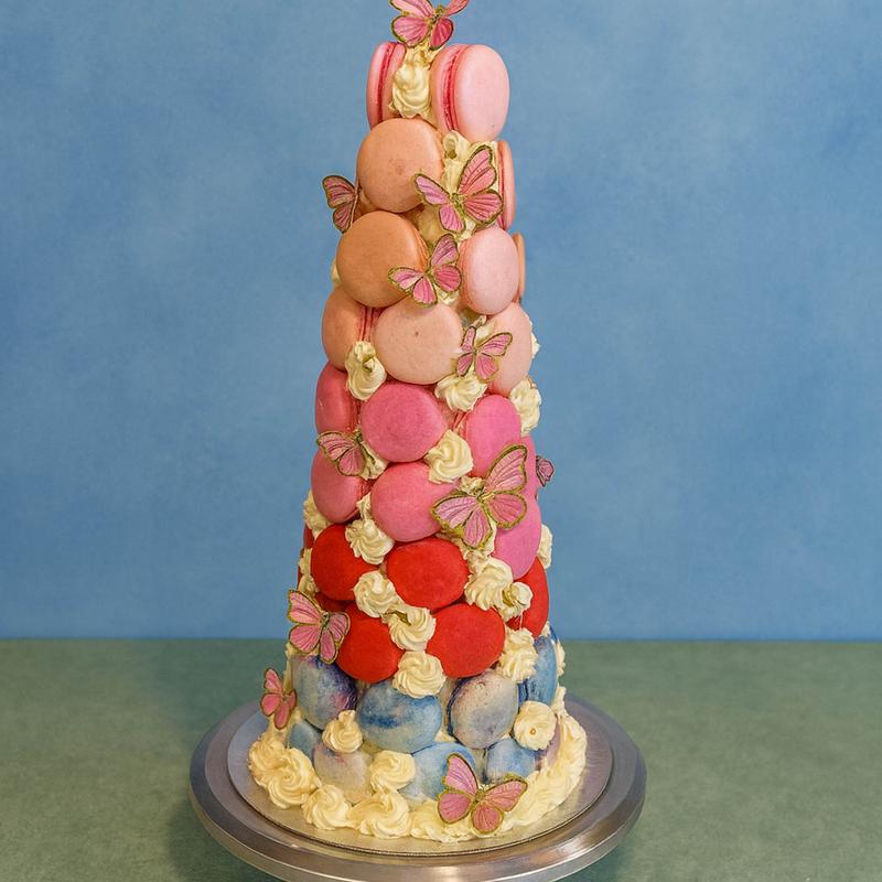 Cone-shaped macaron tower graded from pale blue at the base through red and pink to blush at the tip, with piped cream rosettes between the shells and gold-edged pink butterflies climbing it.