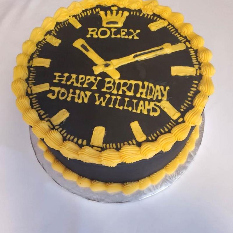 Round cake iced as a watch face in charcoal with yellow piped hour markers, hands and a crown motif, lettered Happy Birthday John Williams, with a yellow shell border.
