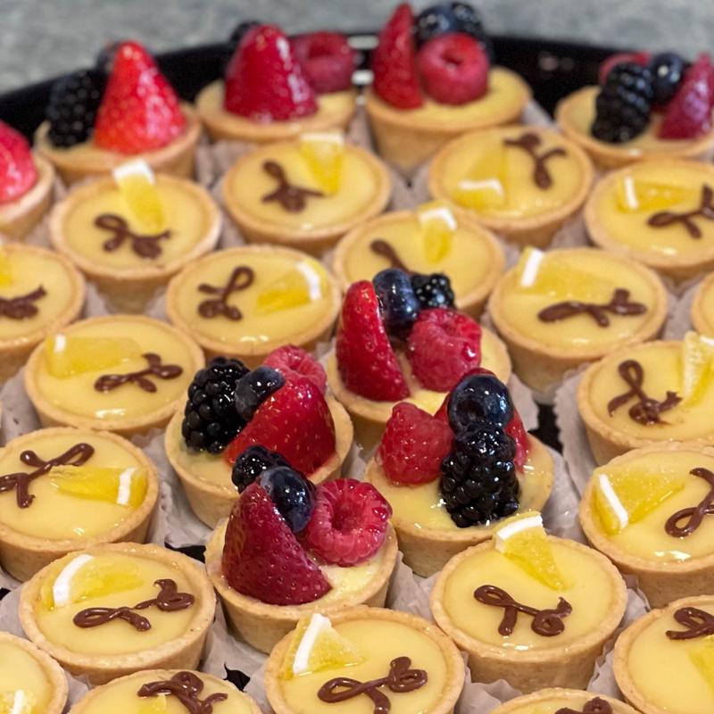 Close-up of a tray of mini tartlettes: pale custard tartlettes finished with a piped chocolate bow and a citrus wedge, interspersed with tartlettes crowned with strawberries, raspberries, blackberries and blueberries.