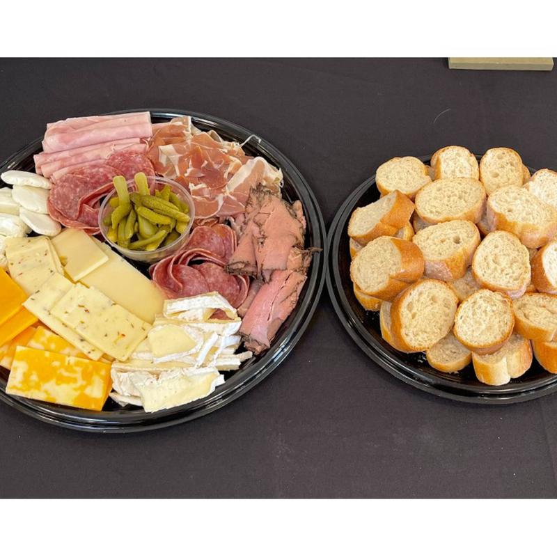 Two black catering platters: one arranged with sliced ham, salami, prosciutto, roast beef, cornichons and an assortment of cheeses, the other filled with sliced baguette rounds.