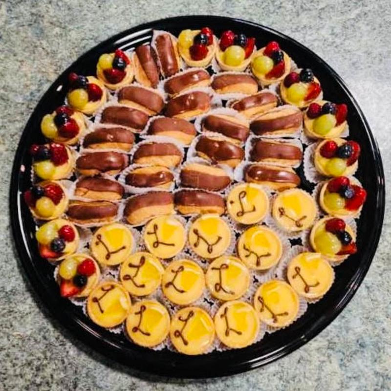 Round platter arranged in rings of chocolate-glazed eclairs, citrus tartlettes piped with a chocolate bow, and fruit tartlettes topped with berries and grapes.