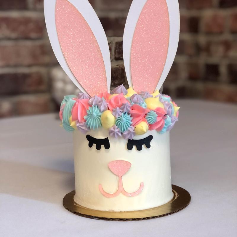 Tall ivory buttercream cake decorated as a sleeping bunny, with black fondant closed eyes, a pink muzzle, a crown of pastel rosettes and meringue kisses, and two glittered pink-and-white ears standing above it.