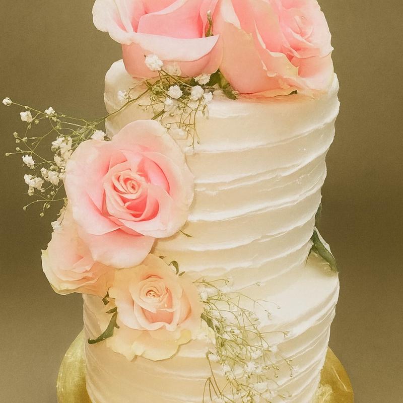 Two-tier wedding cake in horizontally combed white buttercream, dressed with fresh pink roses and sprays of baby's breath cascading down one side.