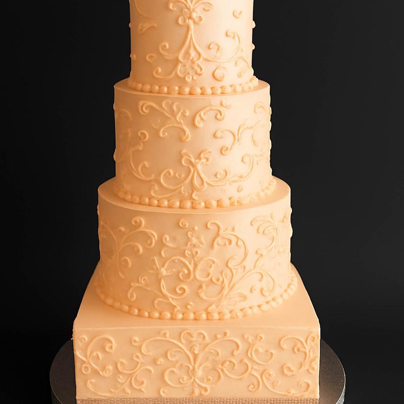 Tiered fondant cake in soft apricot, each tier piped with matching scrollwork and edged with a pearl border, the square base tier finished with a hessian band.