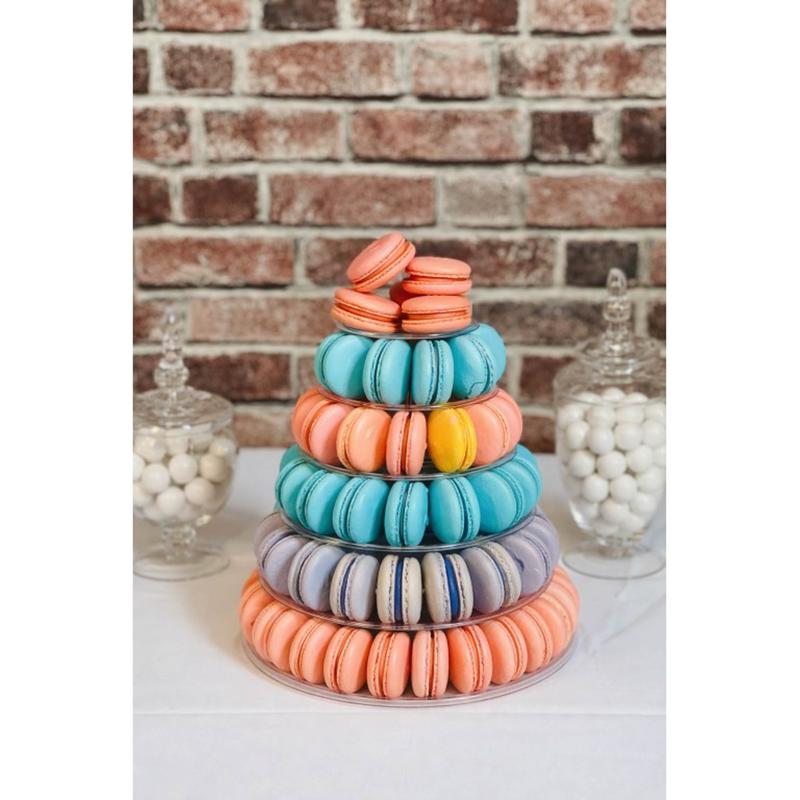 Tiered acrylic macaron tower stacked with coral, turquoise, lavender and cream macarons, flanked by glass apothecary jars of white candy against a brick wall.
