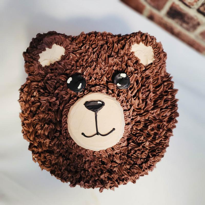 Cake carved and piped as a bear's head, the fur textured in chocolate buttercream with a pale muzzle and inner ears and black icing eyes and nose.