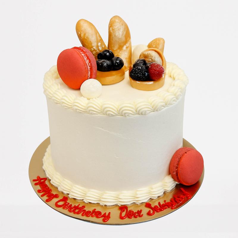 Ivory buttercream cake with piped shell borders, topped with miniature baguettes, tartlettes filled with berries and a coral macaron, and birthday lettering piped in red on the gold board.