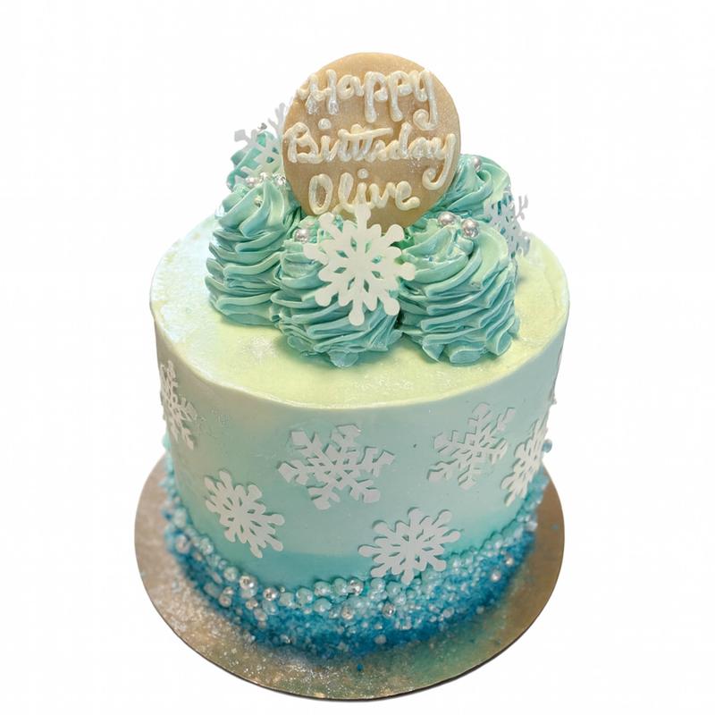 Winter birthday cake in mint-to-blue ombre buttercream, dressed with white fondant snowflakes, a band of blue sugar crystals around the base, ruffled rosettes on top and a round plaque reading Happy Birthday Olive.