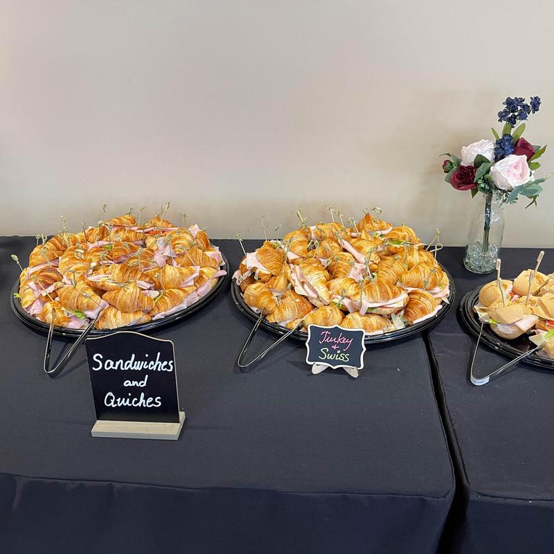 Catering table with two black platters of mini croissant sandwiches on cocktail picks, chalkboard signs reading Sandwiches and Quiches and Turkey and Swiss, and a jar of roses beside them.