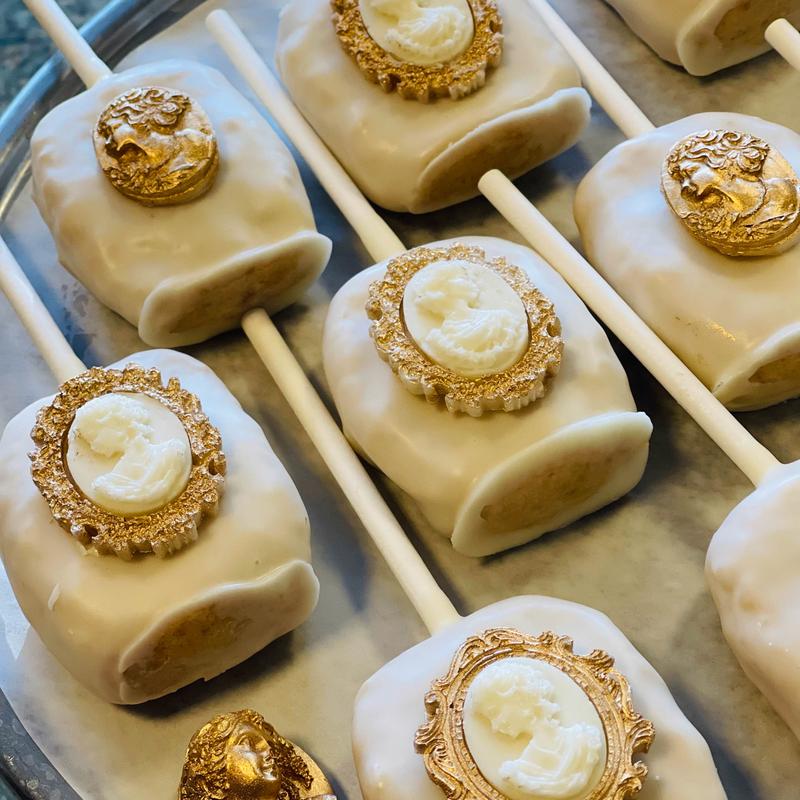 White-glazed cake pops on sticks, each set with an ornate gold-framed cameo medallion, on a silver tray.