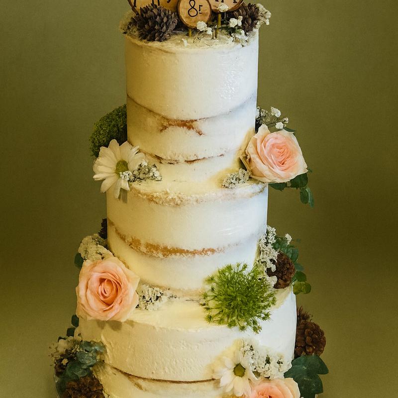 Three-tier semi-naked wedding cake against an olive backdrop, the sponge showing through a thin white coat and dressed with blush roses, white daisies, thistle, pinecones and moss.