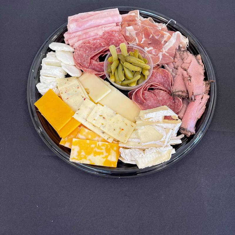 Round black catering platter arranged with rolled ham, salami, prosciutto and roast beef, a cup of cornichons, and a fan of cheddar, pepper jack, swiss and brie.