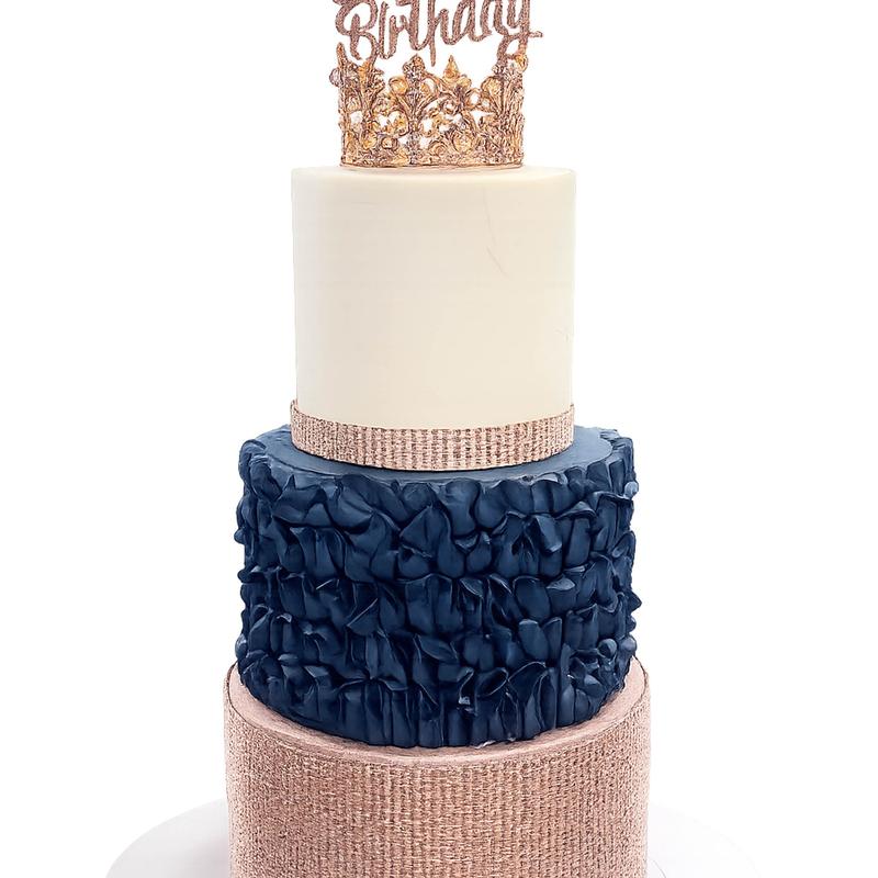 Three-tier cake with a rose-gold sequinned base, a navy ruffled middle tier and a smooth ivory top tier, finished with a gold jewelled crown and a glitter Birthday topper.
