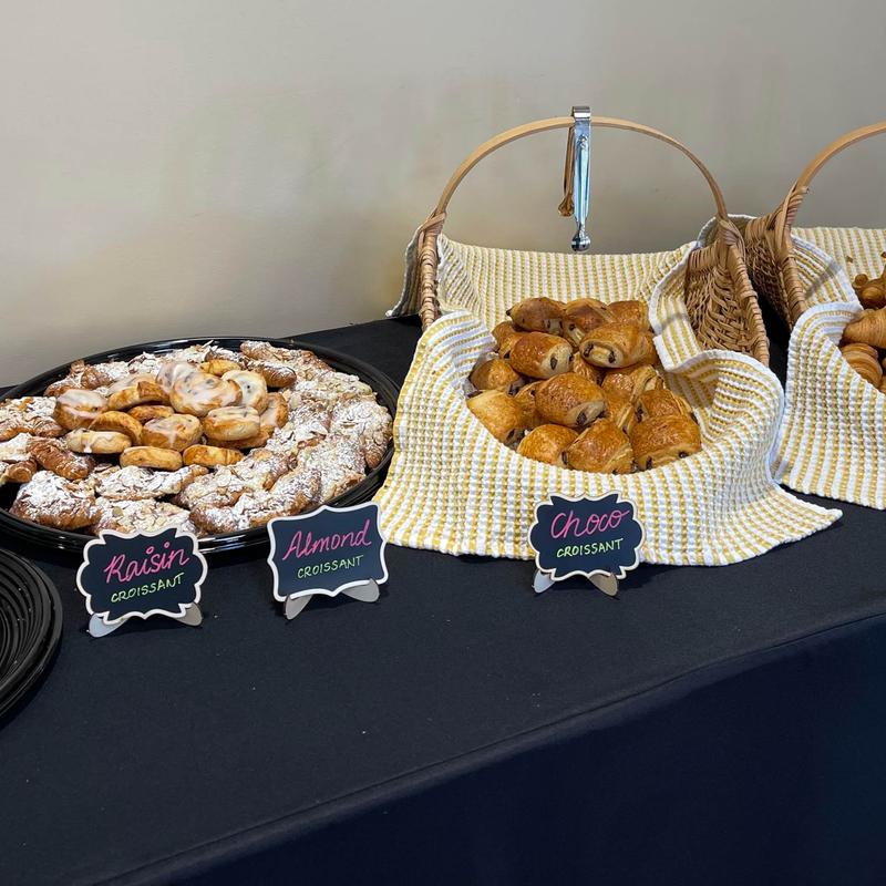 Catering table laid with a platter of raisin and almond croissants beside cloth-lined baskets of chocolate croissants, each labelled with a small chalkboard sign.