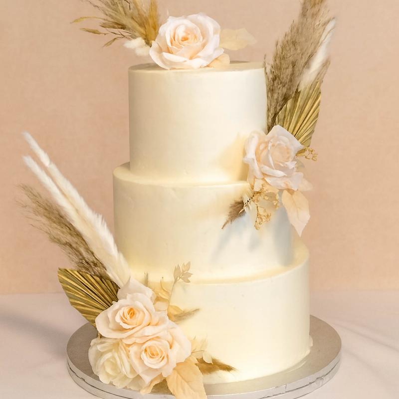Three-tier ivory buttercream wedding cake in a boho dressing of cream roses, dried palm fans, pampas grass and gilded leaves.
