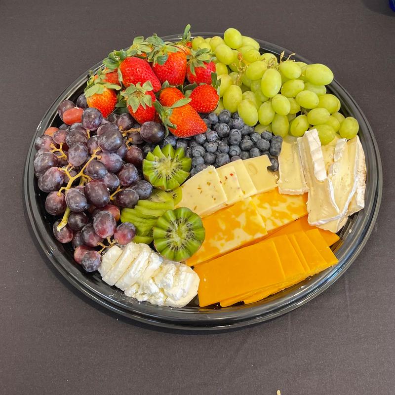 Round catering platter of fruit and cheese seen from above: strawberries, red and green grapes, blueberries and kiwi beside wedges of brie, cheddar and pepper jack.