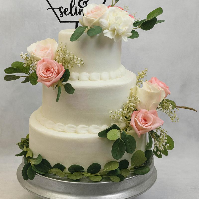 Three-tier ivory buttercream wedding cake with pearl piping between the tiers, fresh pink and cream roses with baby's breath, and a eucalyptus garland circling the base.