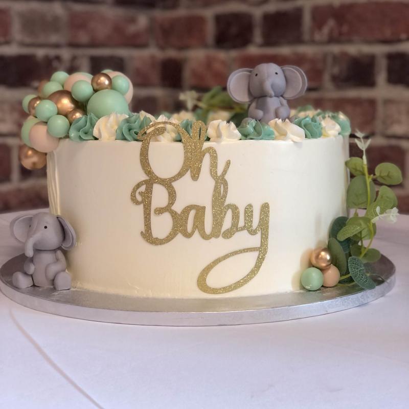 Ivory buttercream cake against a brick wall, with a green and cream rosette border, grey fondant elephants, a cluster of mint, peach and gold balloons and a gold glitter Oh Baby cutout.