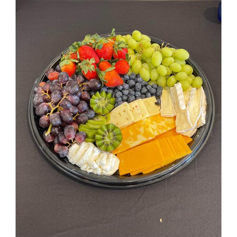 Round catering platter of fruit and cheese seen from above: strawberries, red and green grapes, blueberries and kiwi beside wedges of brie, cheddar and pepper jack.