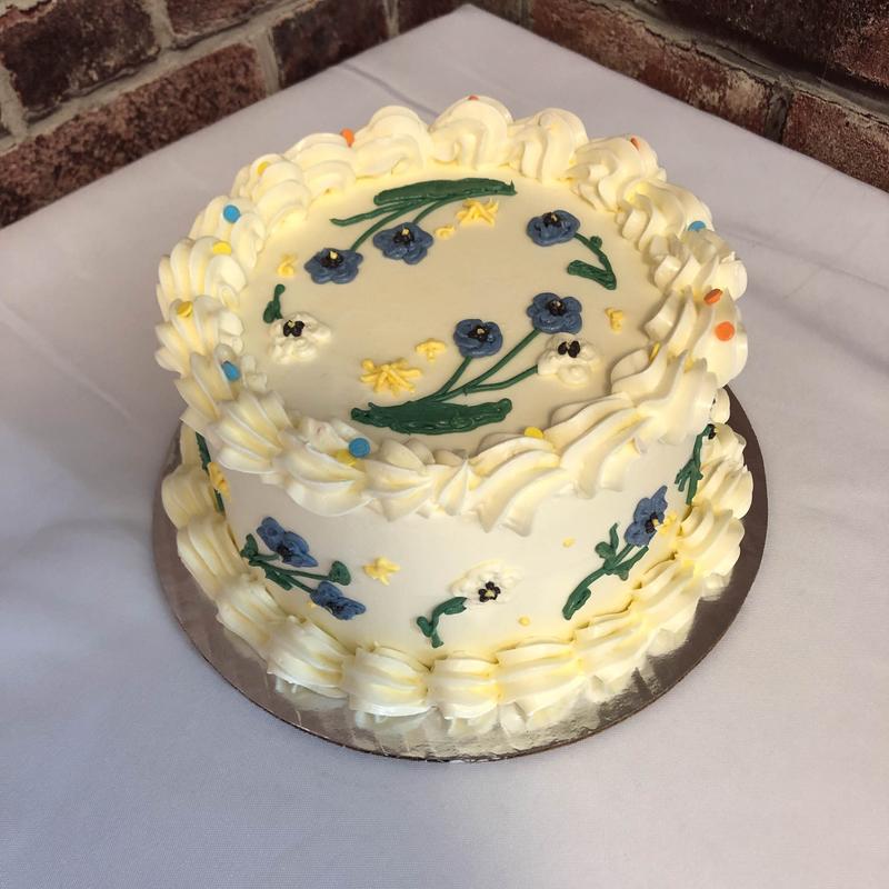 Two-tier ivory buttercream cake with piped shell borders and hand-painted blue and white flowers on green stems, scattered with coloured confetti dots.